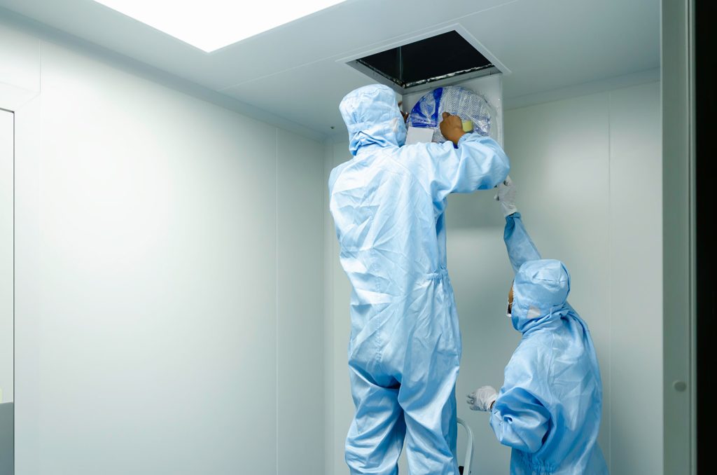 Two technicians wearing full blue protective cleanroom suits, hoods, and gloves perform maintenance in a sterile white room. One worker stands on a ladder reaching up into an open square panel in the ceiling to install or inspect a circular component, while the second worker stands below looking up and assisting.